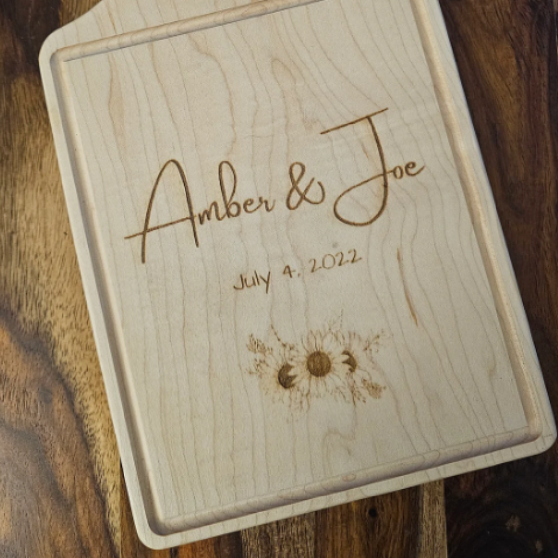 Wooden cutting board with engraved names 'Amber & Joe' and date 'July 4, 2022' on a wooden surface.