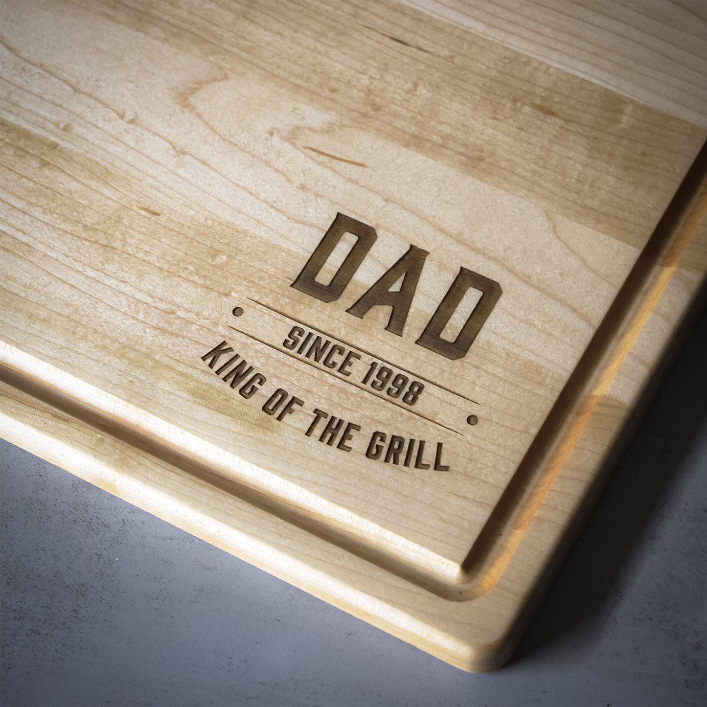 Wooden cutting board with engraved text 'DAD SINCE 1998 KING OF THE GRILL' on a gray surface.