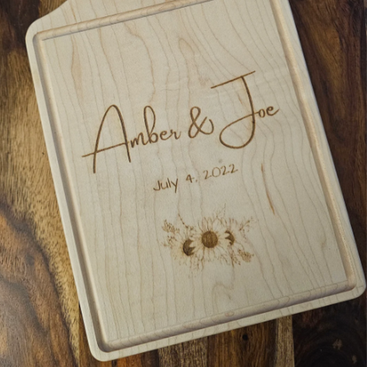Wooden cutting board with engraved names 'Amber & Joe' and date 'July 4, 2022' on a wooden surface.