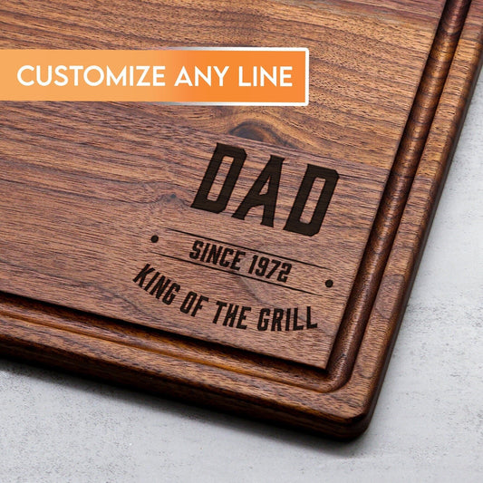 Wooden cutting board with engraved text 'DAD SINCE 1972 KING OF THE GRILL' on a gray surface.